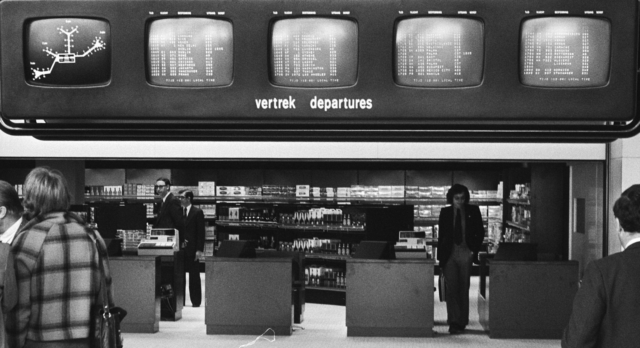 Black and-white photo from the 1970s of businessmen in an airport walking underneath a bank of monitors labeled 'vertrek / departures'