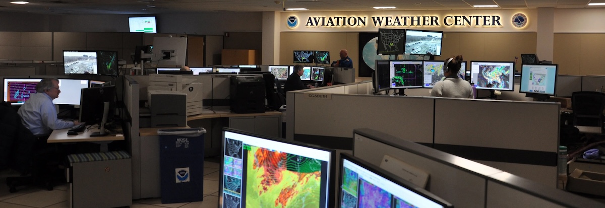 A 2018 photograph of the Aviation Weather Center office interior. Many people working hard at many workstations, all showing colorful and interesting maps of weather phenomena over the US. Image borrowed from https://inspire.eaa.org/2018/03/21/aviation-weather-evolving-for-the-future/.
