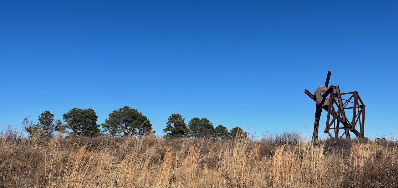 A photograph of a meadow under a cloudless blue sky. Some trees are in the distance. Closer and to one side is a curious metal sculpture, resembling a set of framework made of tumbled-together metal beams.