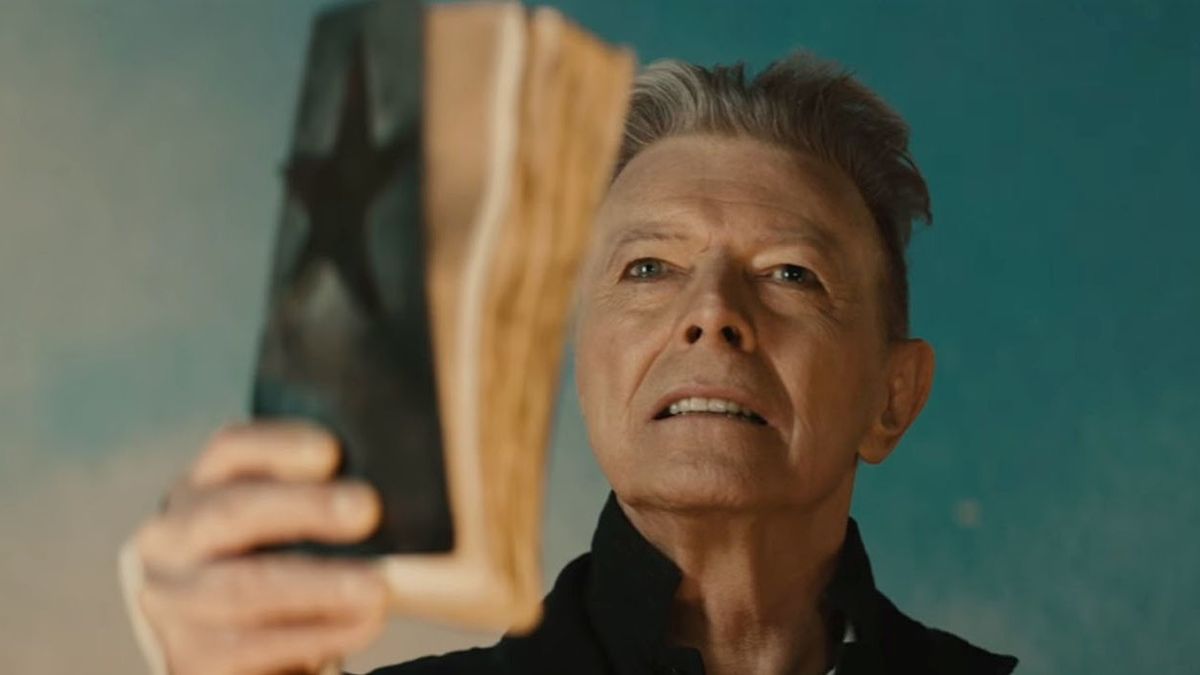 A still image of an older David Bowie holding a tattered book with a black star on its cover.