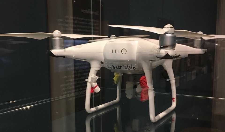 Photograph of a small, white, quad-propellor drone within a dimly lit glass case in a museum. 'Water is Life' is written in black ink on one side, and colored cloth is tied to its four legs: black, red, white, yellow, respectively.