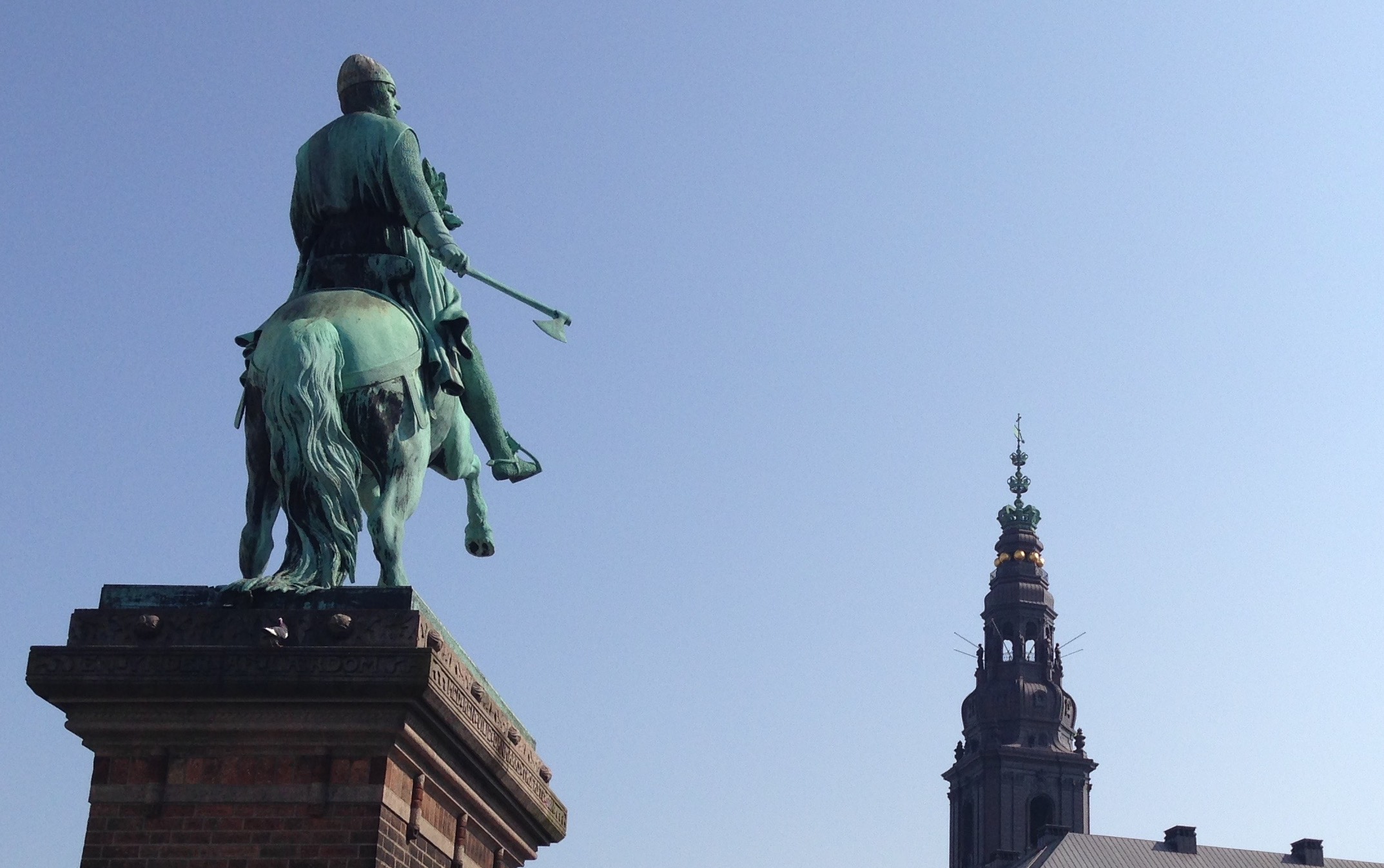 Photograph of a statue of a mounted, armored man holding an axe, and looking at a steeple in the near distance.