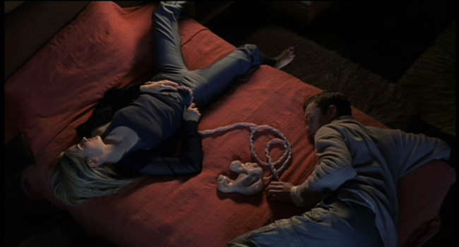 A dimly lit film still depicting a woman and a man reclining on a bed, fully clothed, connected via umbilical cords to a strange, fleshy object lying between them.