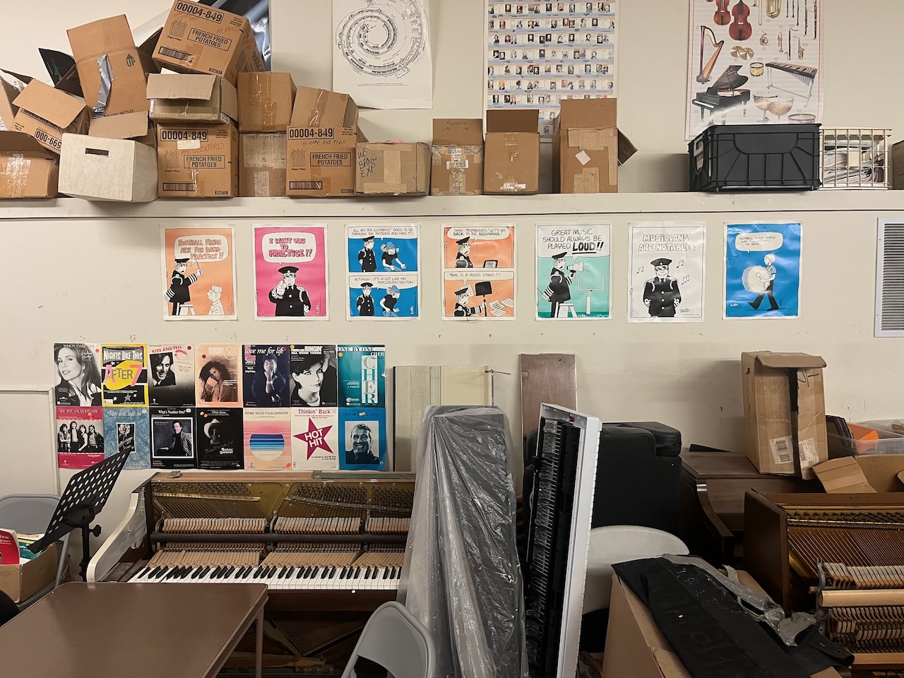 A collection of cartoon posters intended for a circa-1980 school band practice room, hanging over some upright pianos and keyboards in various states of disrepair.