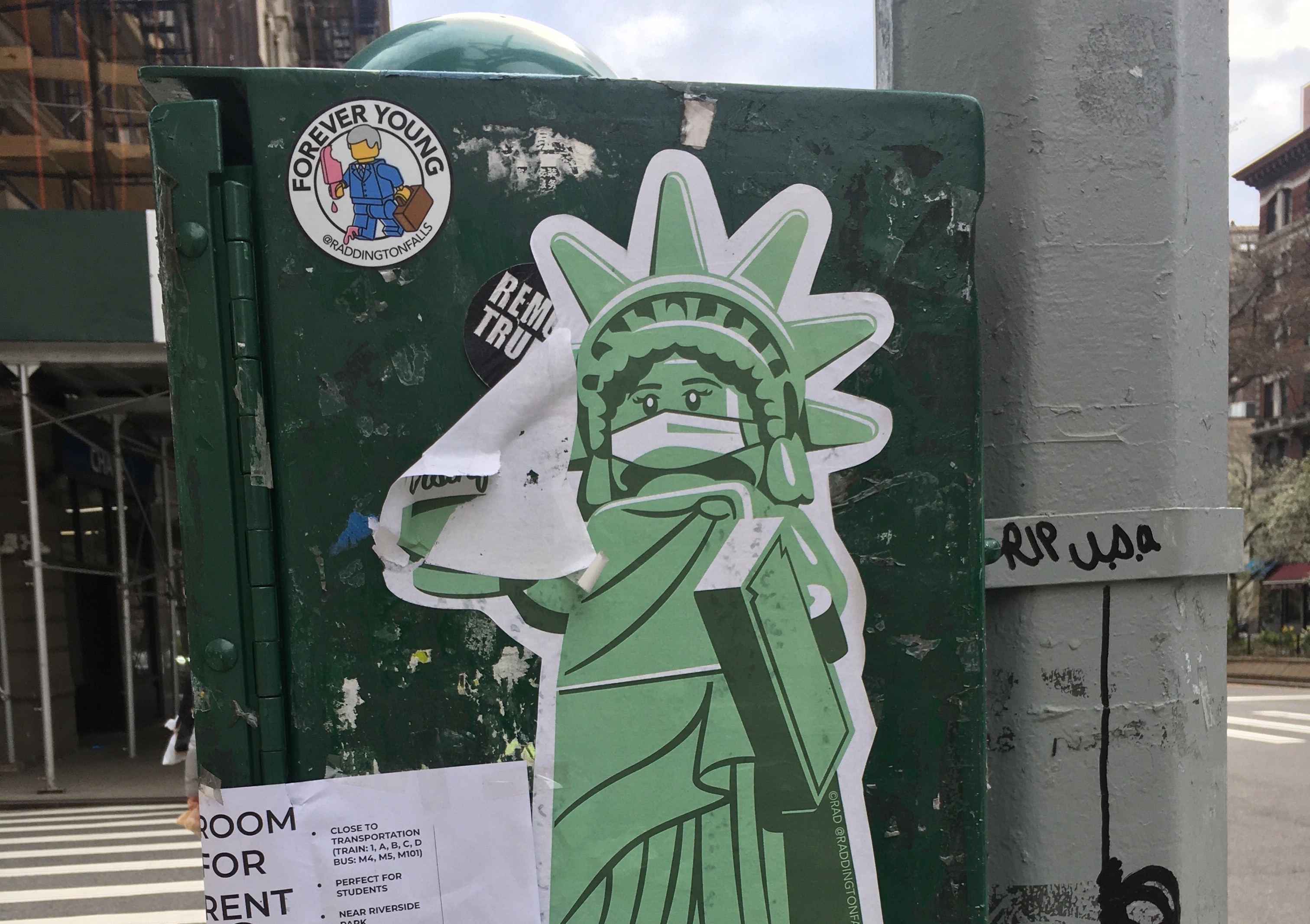 Photograph of several stickers on a utility pole, the most prominent of which depicts the Statue of Liberty wearing a surgical face-mask.