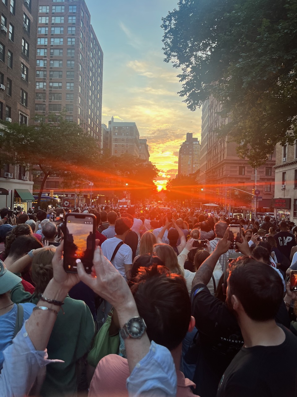 Standing in a crowd in the middle of 79th Street in Manhattan, looking west, towards the setting sun. Rays of brilliant red light spill between buildings, trees, and low, distant clouds. Lots of people are also holding up their phone-cameras to capture this annual spectacle.