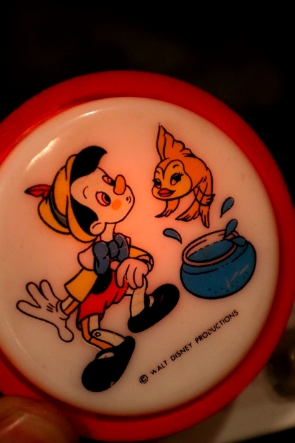 A small, round piece of plastic with a simple, colored drawing of Disney's Pinocchio looking at Cleo the goldfish leaping from her bowl. The top part of it is glowing very softly.
