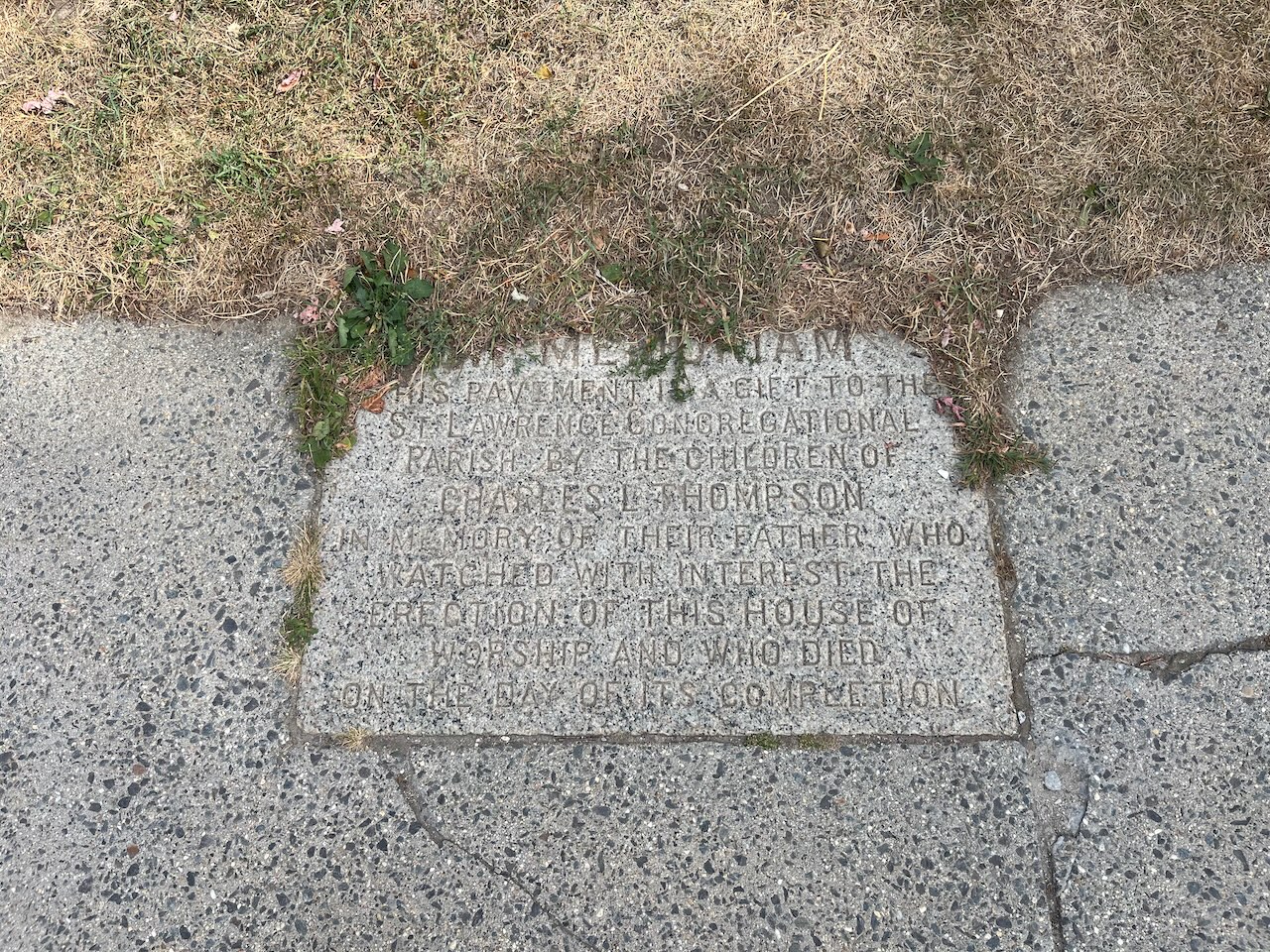 An age-worn pavement stone set into a sidewalk, weeds blooming around its borders. The text is in the attached post.