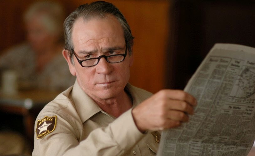 Tommy Lee Jones as a sheriff, holding a newspaper, offering side-eye to some target off-camera.